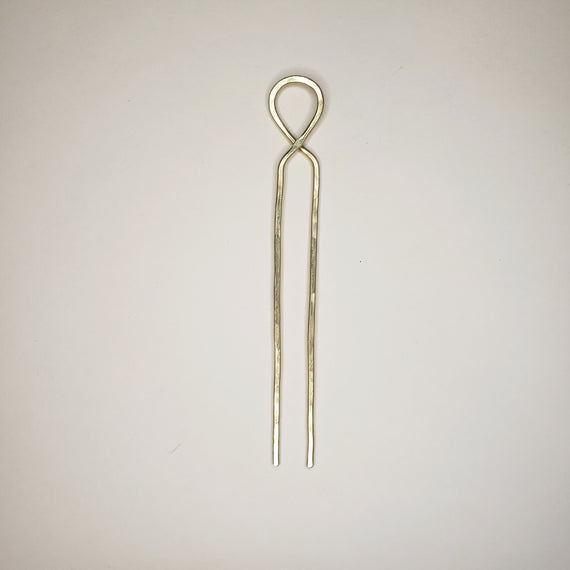 hand hammered brass hairpin laying on a neutral background the hairpin features a crossover loop at the top and two straight prongs