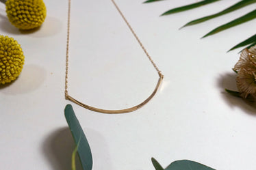 Gold necklace laying flat on white background with leaves. The necklace shows a fine gold chain with a metal shallow half circle shaped hammered bar in the middle of the chain. 