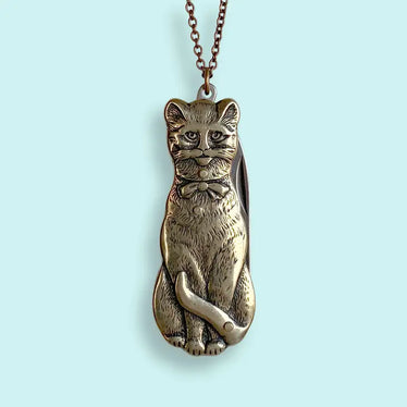 close up shot of cat knife pendant. bronze cat shows carved detailing of sitting cat with tail wrapping around body. the cat has a sullen expression and is wearing a bow. a knife can fold out of the cat. 