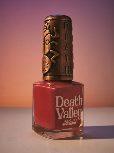 Nail polish bottle sitting on sunset toned background. The bottle has a decorative bronze cap with a beetle sitting on top of a ball and the words death valley. 