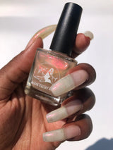 dark skin toned hand holding nail polish bottle and polish swatched on nails. the color looks bright and reflective, with a beautiful peachy shimmer