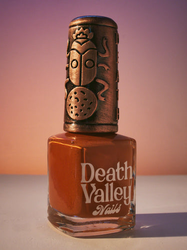 Nail polish bottle with sunset toned background with bronze decorative cap showing a beetle sitting on a rock