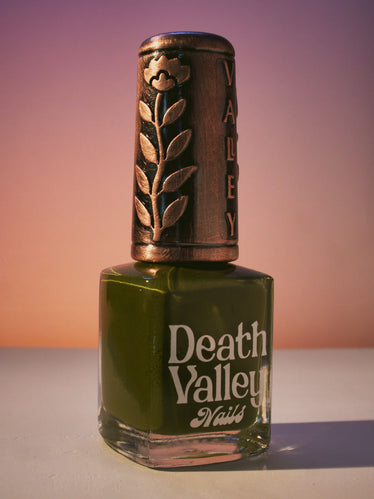 Nail polish bottle on Sunset toned background shows a bronze decorative cap with engraved flower