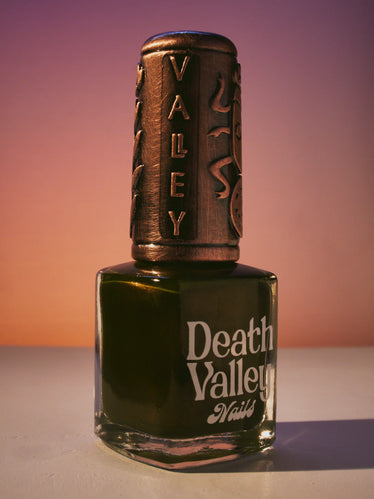 Nail polish bottle on background of sunset towns with a decorative bronze cap featuring the word is death Valley and a beetle sitting on a ball