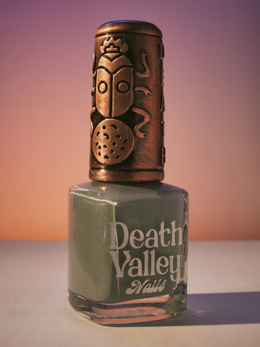 nail polish bottle in Sunset toned background with decorative bronze cap featuring a beetle sitting on a ball￼