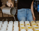 Woman in jeans pouring candle wax from a metal pitcher into frosted glass jars with wicks sticking up. 
