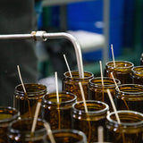 empty amber glass jars lined up with wicks being filled with candle wax by a metal pipe