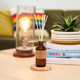 Amber glass bottle with brown paper label with reed sticks poking out on a coffee table next to lamp and plant￼