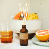 reed diffuser sitting on counter next to fruit bowl. glass of juice. and plate of citrus 
