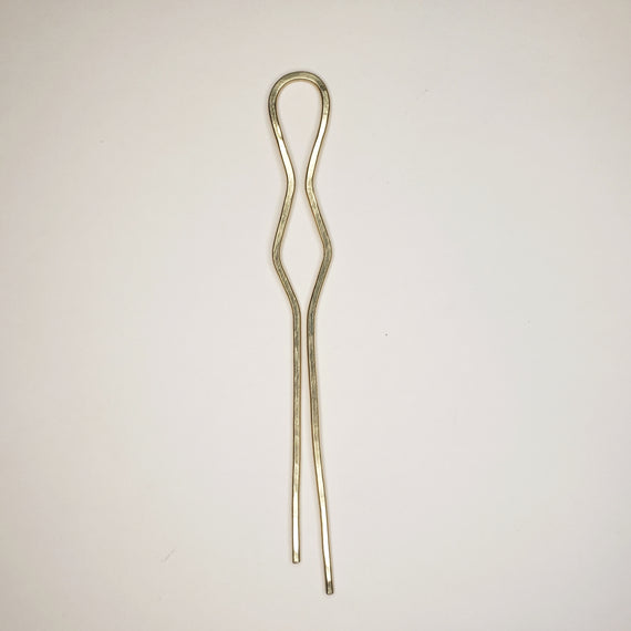 hammered brass hairpin on neutral background.  hairpin features two prongs, a cylindrical loop at the top, and a triangular symmetrical shape in the middle￼