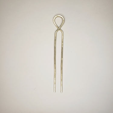 hand hammered brass hairpin laying on a neutral background the hairpin features a crossover loop at the top and two straight prongs￼