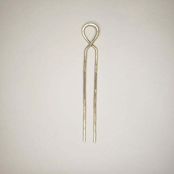 hand hammered brass hairpin laying on a neutral background the hairpin features a crossover loop at the top and two straight prongs￼