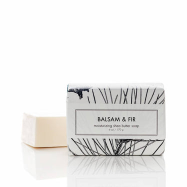 Bar soap wrapped in white paper with decorative black pattern sitting next to unwrapped cream toned bar of soap. 