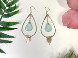 two earring lay on white background. each earring features a golden hammered teardrop shape with 5 golden fringe bars hanging from the bottom and a light teal chalcedony stone hanging within the center of the shape.