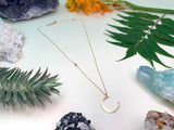Crescent moon necklace laying flat on white background with flowers. shows a delicate gold chain with a cut in labradorite bead and hammered gold crescent moon￼