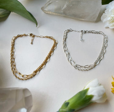 two double chain bracelets laying on white background with flowers one in gold, one in silver￼