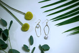 Two earring laying flat on white background with flowers. The earrings are abstract oval shapes. One is longer and has a pearl hanging inside of it and the top by the ear wire. The other is shorter and has a pearl hanging off the bottom. making the two earrings the same length 
