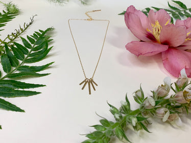 Delicate gold necklace laying on white background with flowers. necklace features five metal sticks which separate one hanging to create a angled fringe