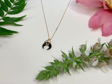 gold necklace laying flat on white background with flowers. the necklaces is a fine gold chain with a wire wrapped crescent moon shaped black horn and small greenish blue clear stone. 