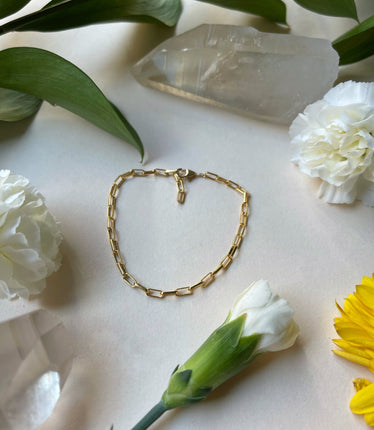 gold bracelet laying flat on a white background with flowers. the gold chain is made of small ocular rectangles linked together, the clasp is latched into the end of the chain 