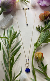 Golden necklace laying flat featuring a rounded lapis stone hanging off of a gold hammered circle connected to the chain with two small lapis beads