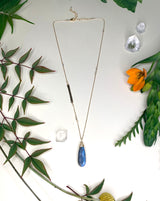 Gold necklace laying flat on white background with leaves. Necklace features a labradorite teardrop pendant, Labradorite beads cut into the chain, and a hammered metal bar cut into the chain. 