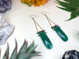 two earrings laying flat on white background. two rounded rectangle shaped green onyx crystals hang by two strands of delicate golden chain. tiny pyrite beads are strung between the two chains. 
