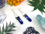 two earrings laying flat on white background. two rounded rectangle shaped lapis stones hang by two strands of delicate golden chain. tiny pyrite beads are strung between the two chains. 
