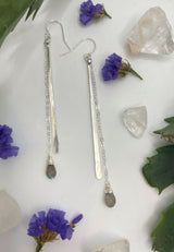 close-up shot of hammered stick with stone drop earrings. earrings consist of one long hammered metal paddle and a length of chain with a labradorite hanging off the end