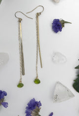 close-up shot of hammered stick with stone drop earrings. earrings consist of one long hammered metal paddle and a length of chain with a peridot hanging off the end