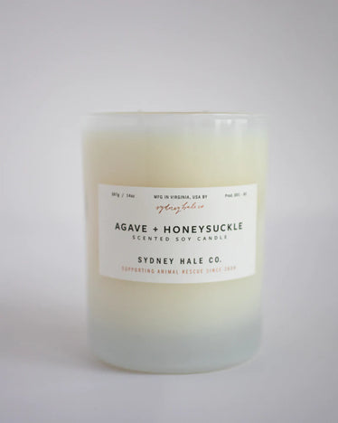 Cream toned candle in white frosted glass with a white label and black writing with red text detailing, sitting on a neutral white background. 