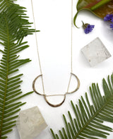 Gold triple arc necklace laying flat on white background with leaves. this necklace features a delicate fine gold chain with three arcs lined up across the chain￼