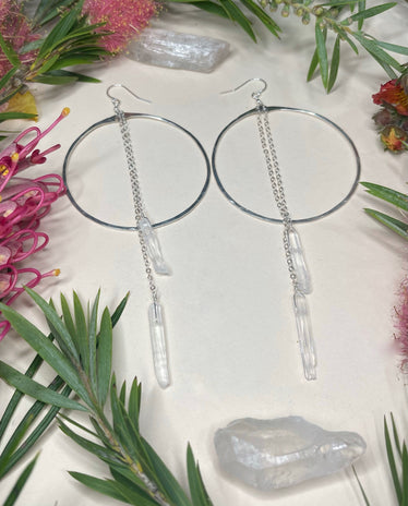 two silver circle hoops laying flat featuring two quartz crystals of different lengths hanging from chains to catch the light￼