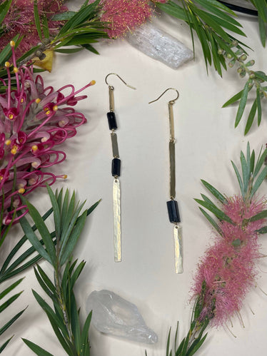 two gold earrings laying flat. one earring has two small rods of black tourmaline separated by three hammered gold bars of the same width. the other earring has three gold hammered bars with one longer black tourmaline rod towards the bottom. 