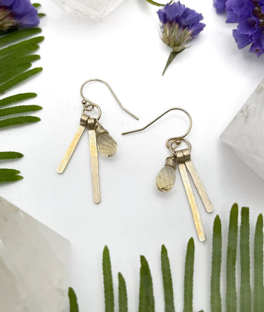 two earrings laying flat on white background each earring features two gold sticks hanging next to a small wire wrapped citrine stone￼