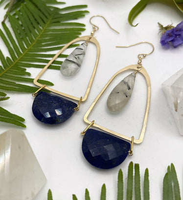 hammered bell earrings on white background surrounded by flowers they show a teardrop tourmalinated quartz stone encapsulated by a metal shape with a lapis half circle hanging off the bottom￼