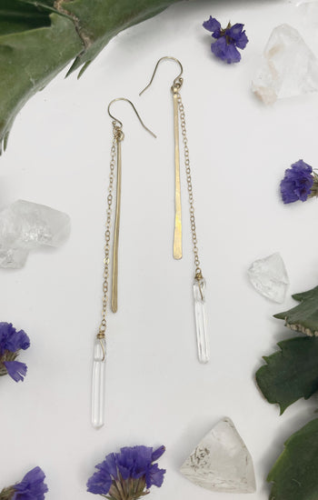 close-up shot of hammered stick with stone drop earrings. earrings consist of one long hammered metal paddle and a length of chain with a clear quartz hanging off the end￼