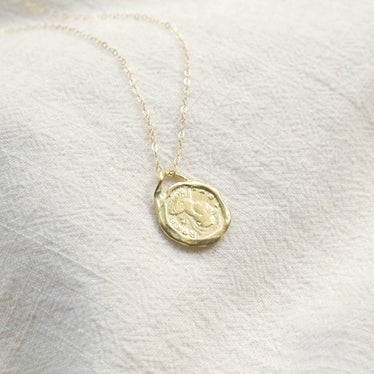 Roma necklace on fabric background.  necklace has gold dainty chain and a round Roman coin stamped pendant